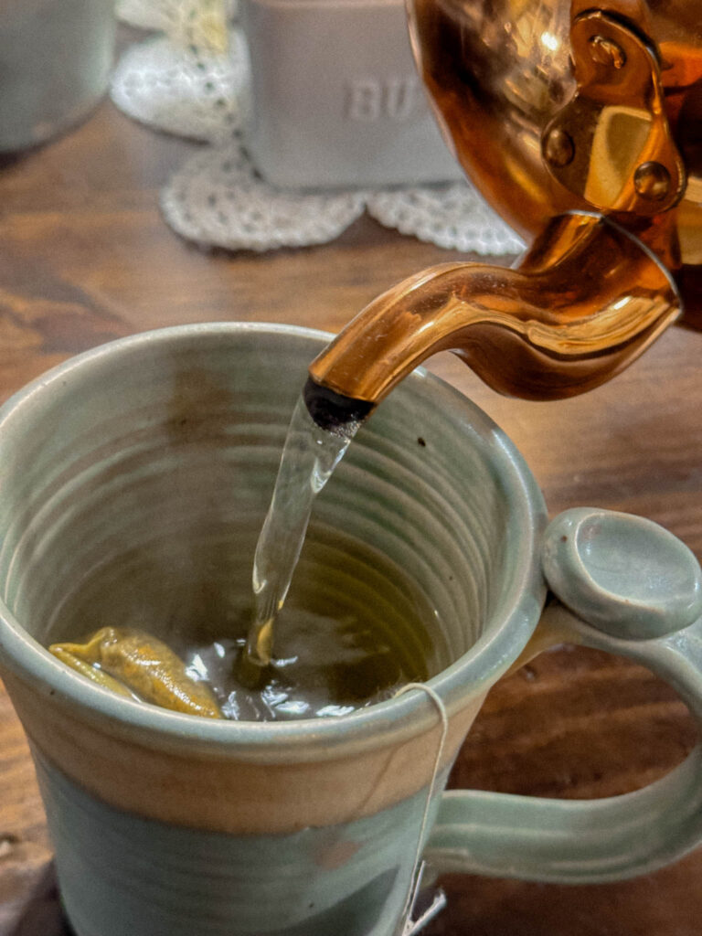 A close-up of hot water being poured from a copper kettle into a green ceramic mug, steeping a tea bag. A lace doily decorate the wooden table underneath.