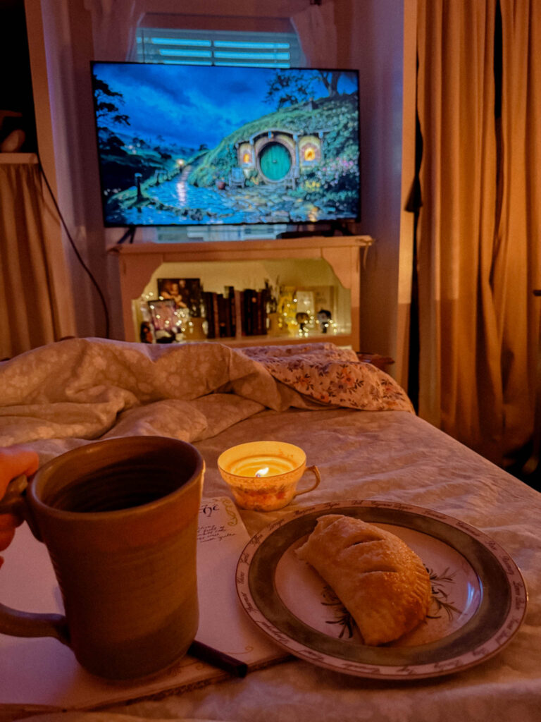 A warm, golden-lit scene with a hobbit hole on TV, a teacup candle, a ceramic mug in hand, a plate with a pastry, and a journal peeking from underneath—all resting on a cozy bed.