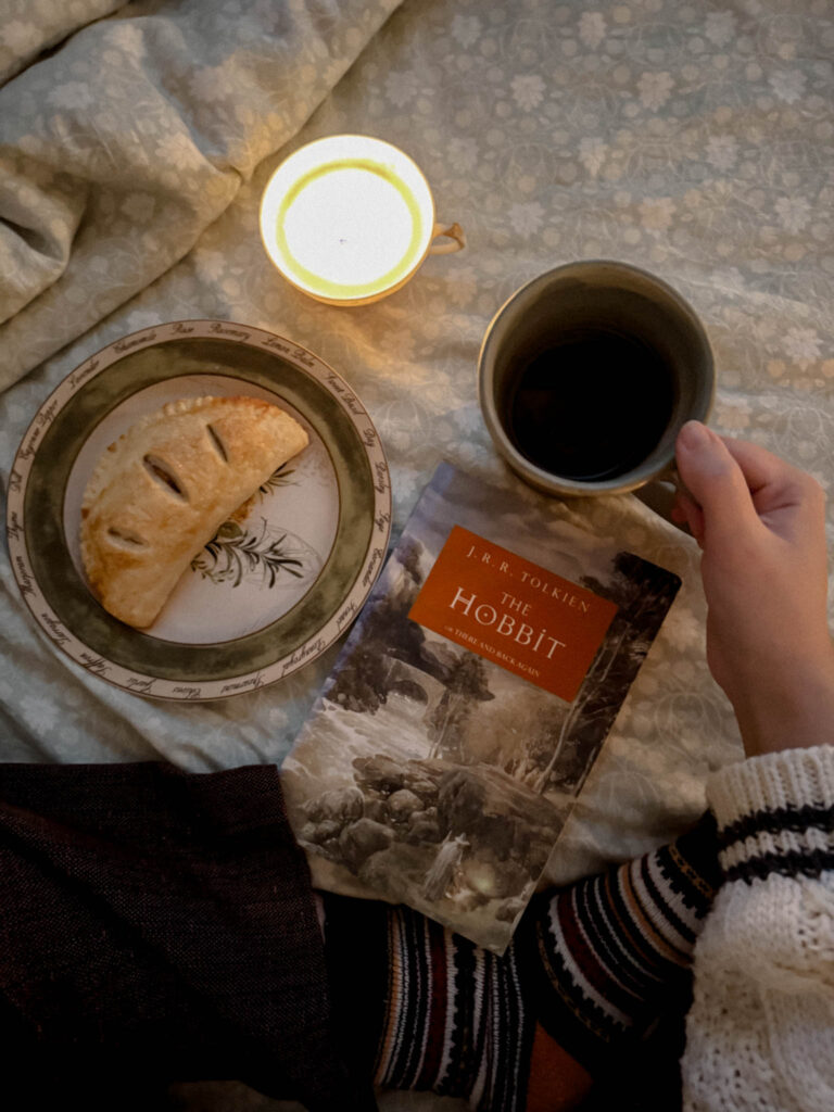A hand holds a copy of The Hobbit next to a plate with a flaky turnover pastry, a teacup candle, and a ceramic mug of black coffee or tea. Cozy socks and soft bedding complete the scene.
