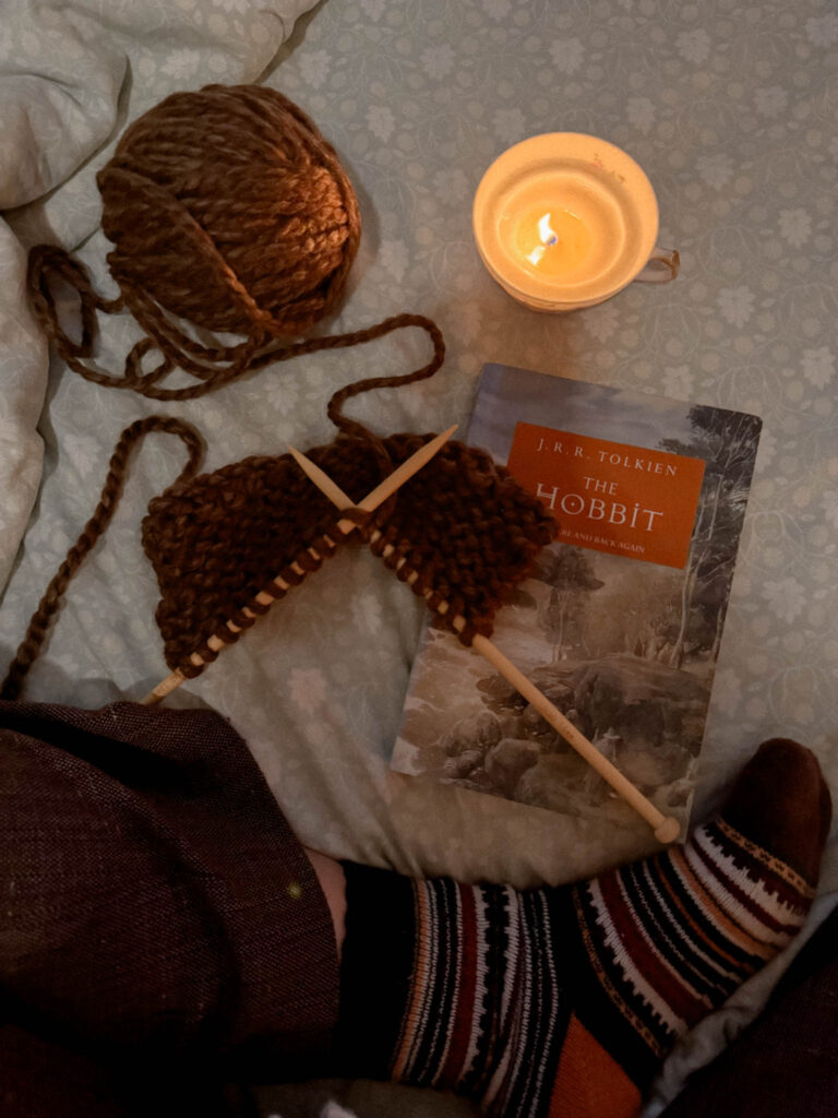 A ball of thick brown yarn sits beside a candle in a teacup, knitting needles mid-project, and a copy of The Hobbit on a soft floral bedspread. A pair of legs in brown pants and striped cozy socks peek into the frame.