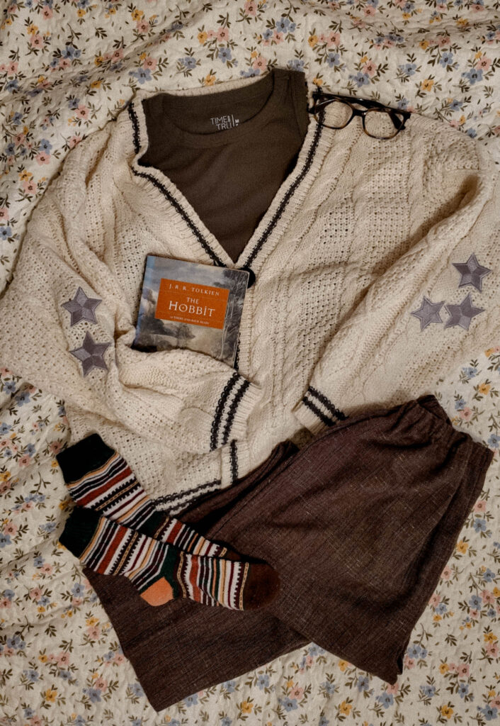 A neatly laid out cozy outfit on a floral bedspread—featuring a cable-knit cream cardigan with star patches, brown lounge pants, a dark green top, striped socks, glasses, and The Hobbit tucked in the cardigan.