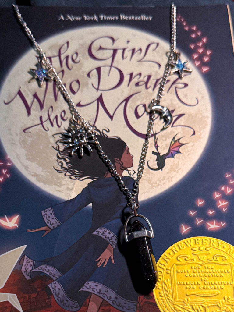 DIY celestial charm necklace with star, sun, moon, and crystal pendant resting on the cover of “The Girl Who Drank the Moon