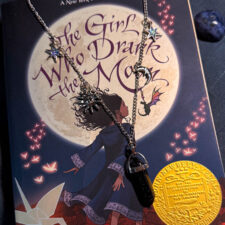 Handmade celestial charm necklace styled on a fantasy book cover, featuring silver moon, star, and sun charms with a crystal point pendant