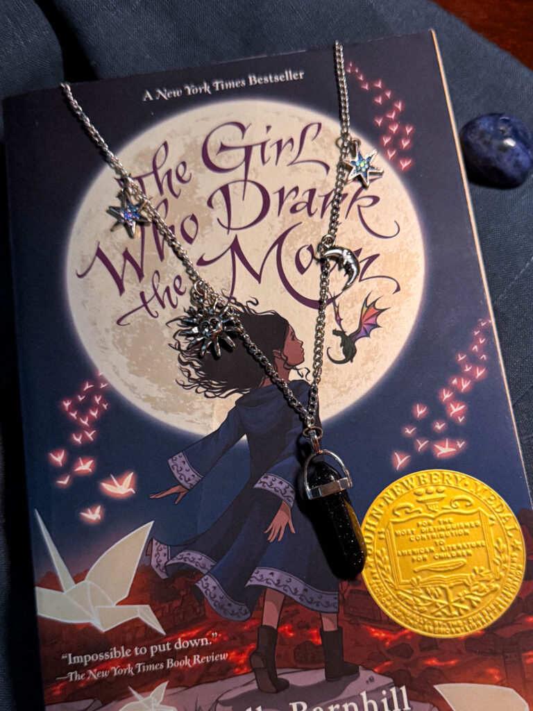 Handmade celestial charm necklace styled on a fantasy book cover, featuring silver moon, star, and sun charms with a crystal point pendant