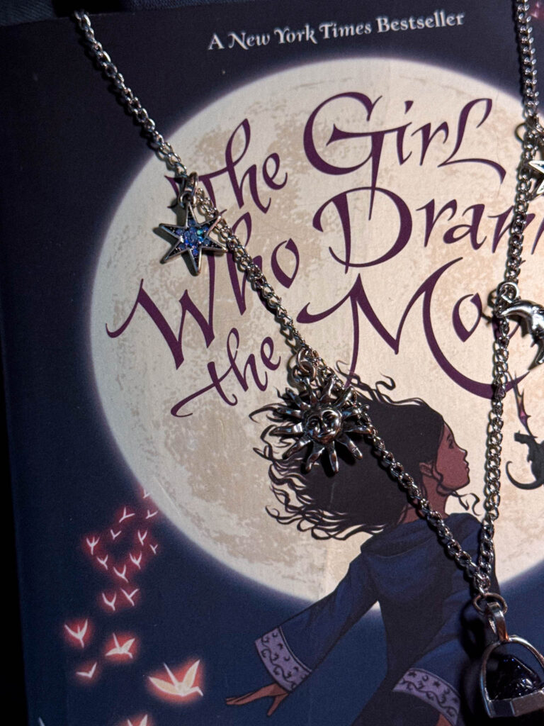 Close-up of a silver sun charm and blue star charms on a celestial necklace draped over the moonlit cover of “The Girl Who Drank the Moon