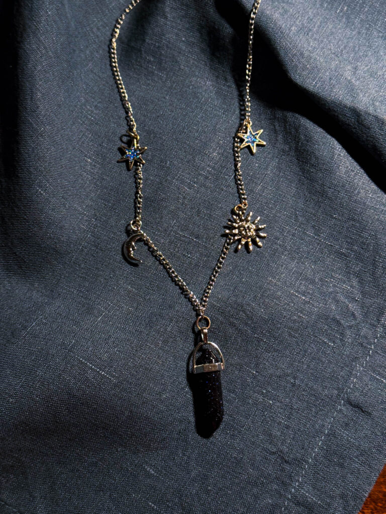 Flat lay of a celestial moon and star necklace with sun charm and crystal pendant on dark linen fabric—cozy DIY styling