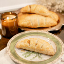 Golden apple hand pie on a floral herb plate, with candlelight and warm pies in the background.