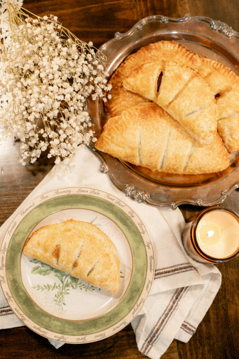 Top-down view of rustic apple hand pies on a silver tray beside dried flowers and a single plated pie.