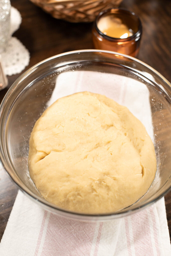 Ball of enriched sweet dough rising in a glass mixing bowl, set on a pink-striped tea towel with warm amber candlelight in the background.