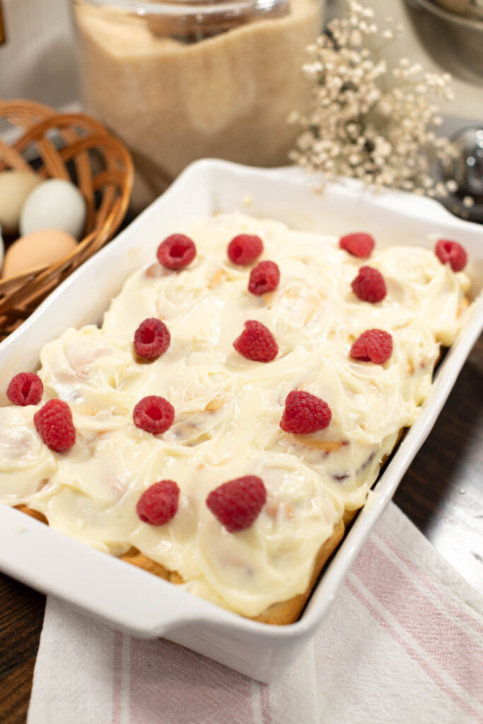 Freshly baked raspberry cinnamon rolls in a white ceramic baking dish, topped with swirls of cream cheese frosting and whole raspberries, ready to serve.