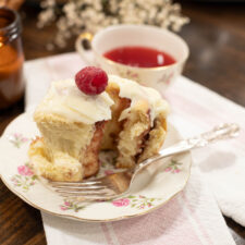 Close-up of a raspberry cinnamon roll with cream cheese frosting and a fresh raspberry on top, served on a vintage floral plate with a silver fork and a cup of raspberry tea.