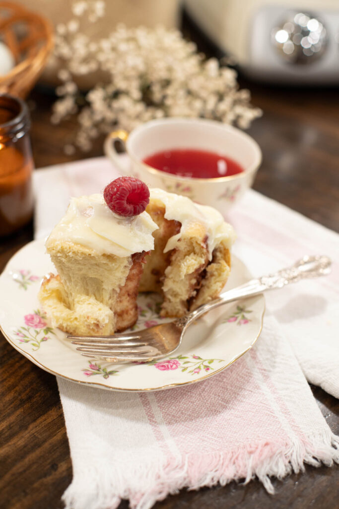 Close-up of a raspberry cinnamon roll with cream cheese frosting and a fresh raspberry on top, served on a vintage floral plate with a silver fork and a cup of raspberry tea.
