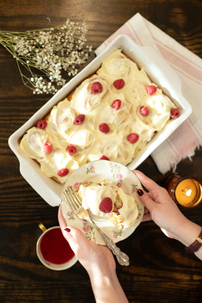 Hands holding a single raspberry roll on a floral plate above a baking dish of frosted rolls, with a cup of tea and soft candlelight on a dark wood table.