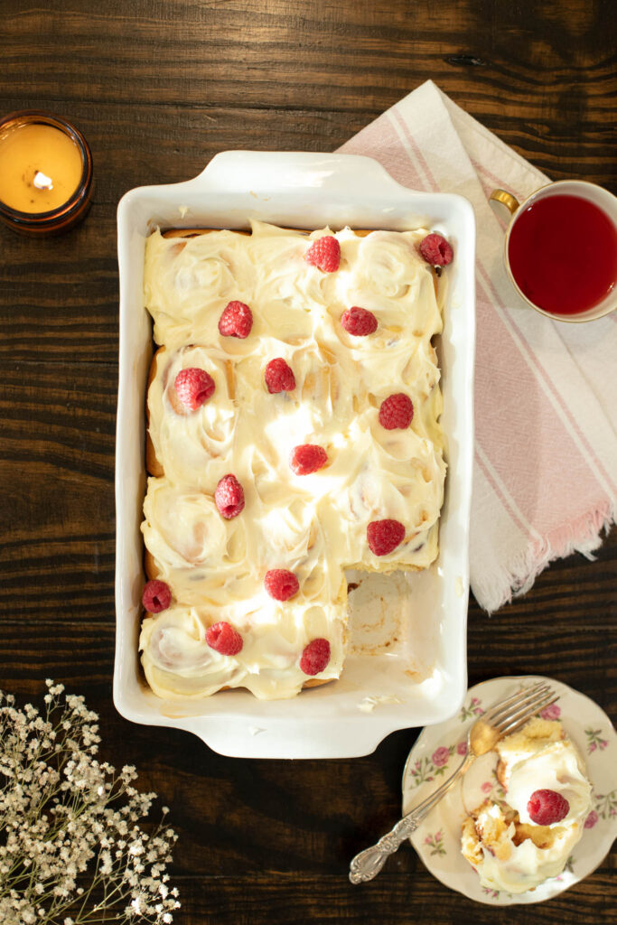 Overhead view of a pan of frosted raspberry cinnamon rolls with fresh raspberries, a missing corner piece, and a tea cup beside a pink striped linen napkin.