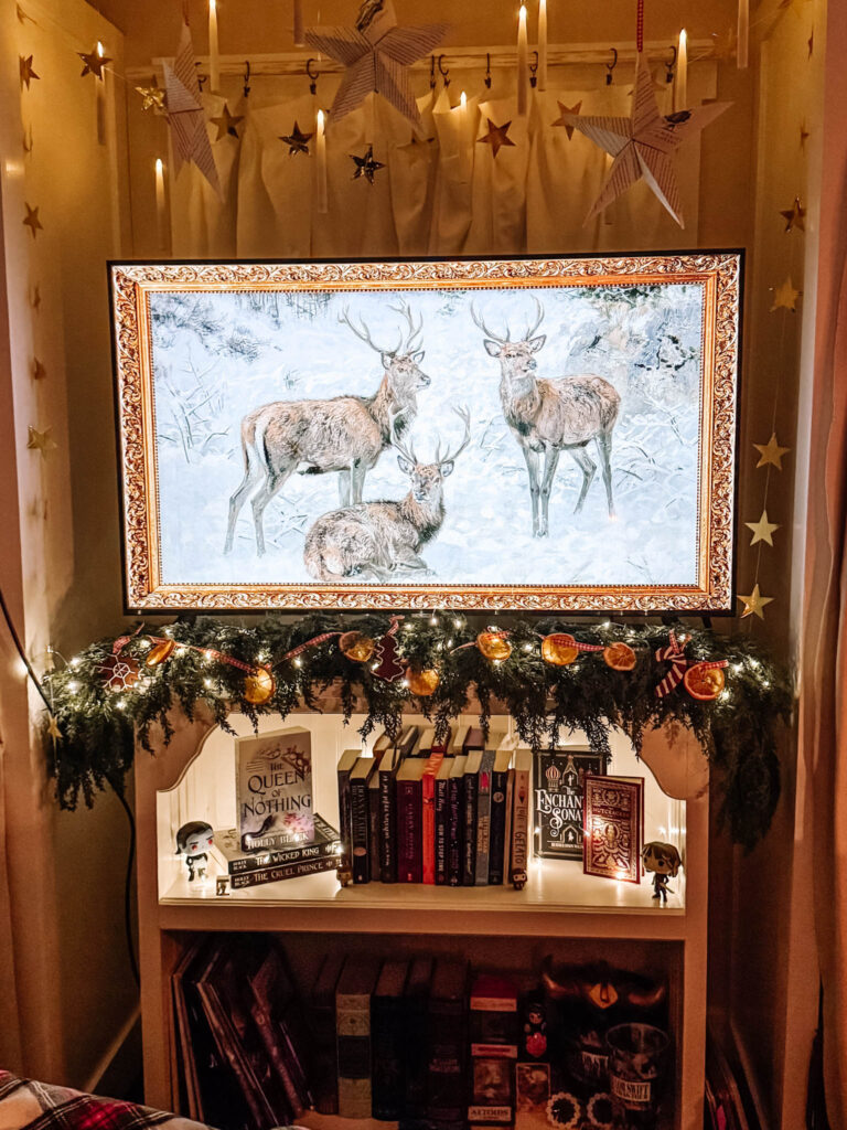 A framed winter scene of three deer displayed on a TV above a white bookshelf. Evergreen garland with dried oranges, gingerbread ornaments, and fairy lights drapes across the shelf, while book series and small figurines fill the cubbies below. Paper stars and hanging candles glow softly in the background.