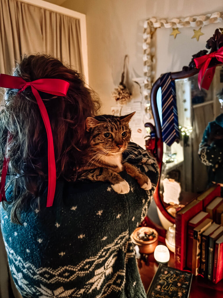 A person with long hair tied in a red ribbon holds a tabby cat over their shoulder. They’re standing beside a vintage vanity decorated with candles, classic books, and cozy holiday garlands. The warm lighting and layered textures create a soft, comforting cottage atmosphere.