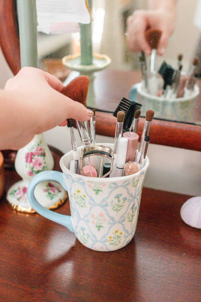 Makeup brushes and everyday beauty items stored in a floral ceramic mug on a wooden vanity, adding a whimsical touch to a morning routine.