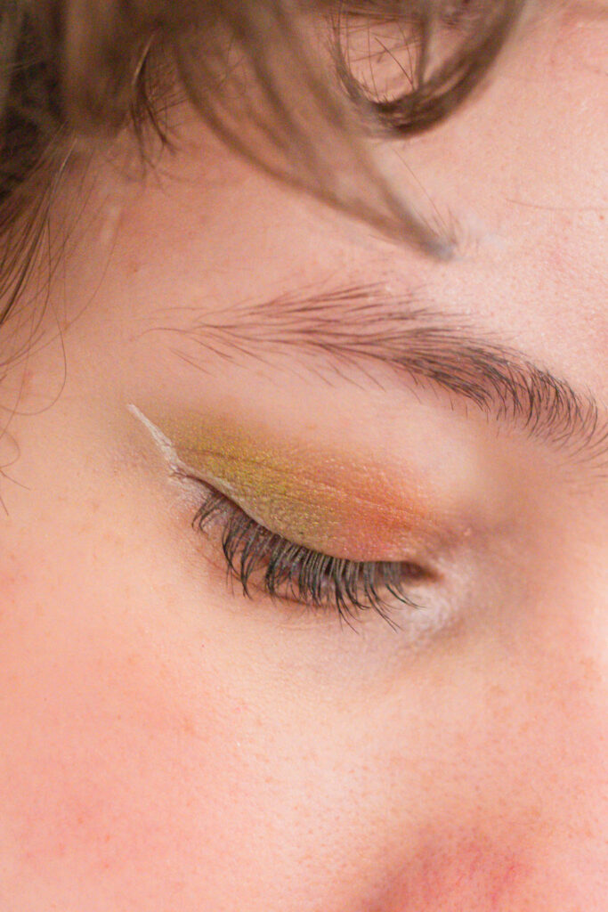 Close-up of a closed eye with soft green and gold shimmer eyeshadow, showing a subtle and playful pop of color.