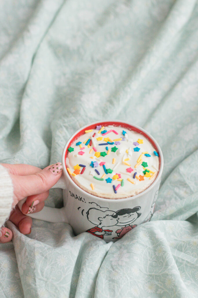 A cozy mug topped with colorful sprinkles held over light blue bedding, capturing a simple moment of everyday whimsy.