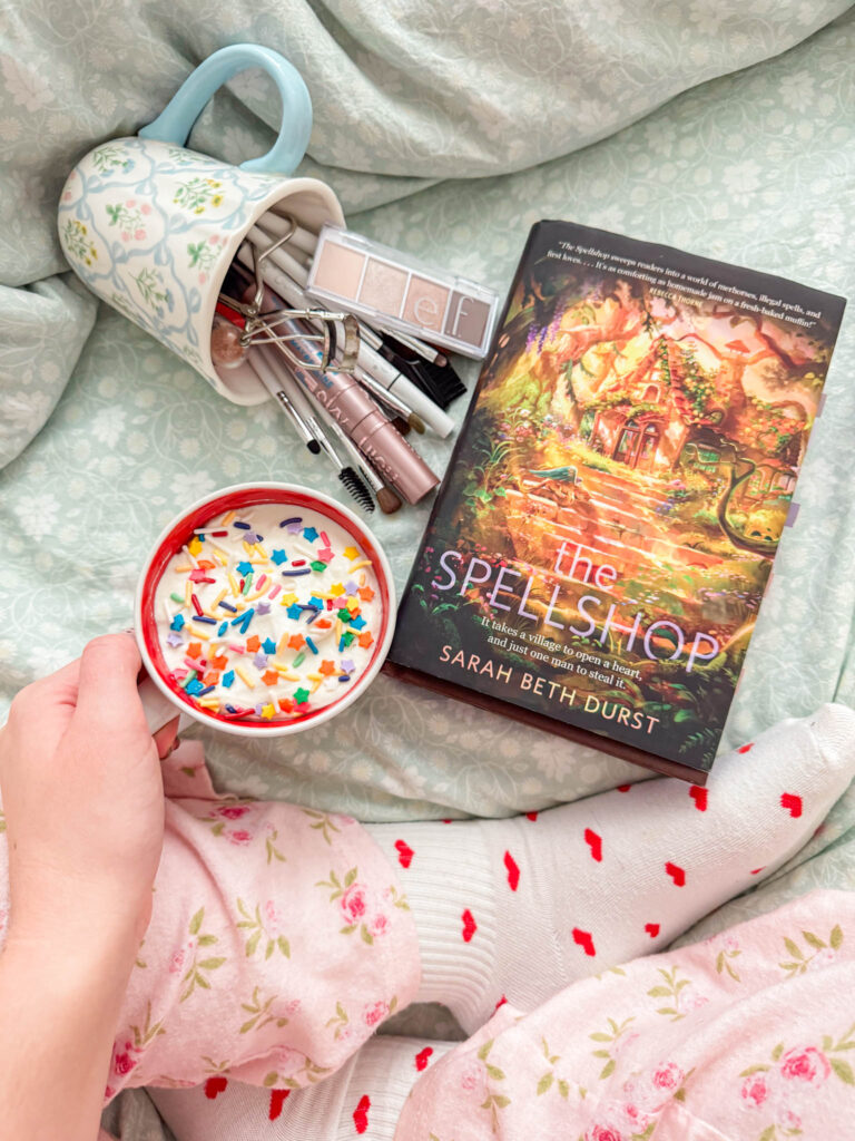 A cozy bed scene with floral pajamas and heart-patterned socks, a sprinkle-topped drink held in hand, a fantasy book resting nearby, and makeup brushes spilling from a floral mug—capturing a quiet moment of everyday whimsy.