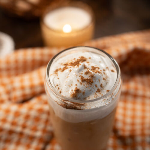 Brown sugar pumpkin iced latte topped with thick vanilla cold foam and a dusting of cinnamon, photographed from above on an orange gingham cloth beside a glowing candle.